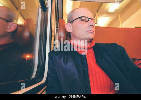 An elderly man in glasses commuting at work. Middle age man looking out of the window of train. Passenger during travel by high speed express train in Europe. An elderly man travelling in train at night Stock Photo