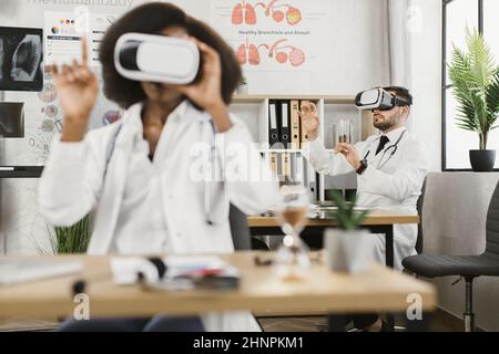 Two multicultural male and female doctors wearing VR goggles while sitting together a medical cabinet. Colleagues getting in air while studying virtual screen. Stock Photo