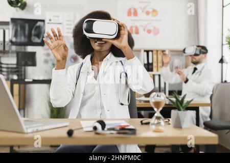 Two multicultural male and female doctors wearing VR goggles while sitting together a medical cabinet. Colleagues getting in air while studying virtual screen. Stock Photo