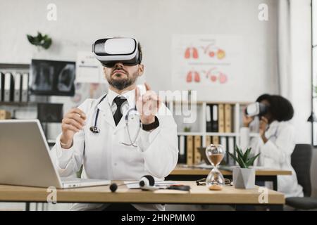 Two diverse medical specialists wearing virtual reality glasses while working at hospital office. Cooperation, innovations and medicine concept. Stock Photo