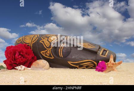 Wooden Tiki Mask on Sand With Flowers and Blue Sky Background Stock Photo