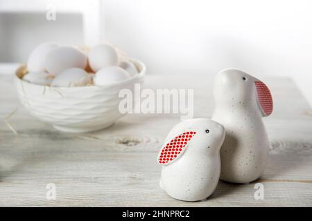 Two figurines of white hares with red ears big and small on a wooden saw cut and a bowl with eggs in the background in a white Scandinavian style kitc Stock Photo