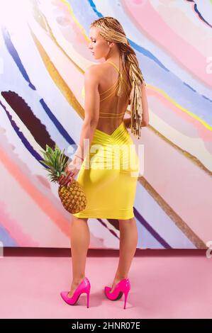 Young, cute, beautiful slim blonde woman with african braids and with  pineapples in her hands in yellow dress on colorful background Stock Photo