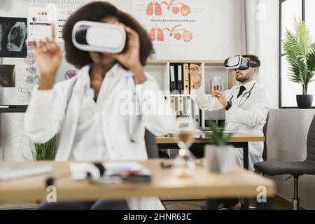 Two multicultural male and female doctors wearing VR goggles while sitting together a medical cabinet. Colleagues getting in air while studying virtual screen. Stock Photo