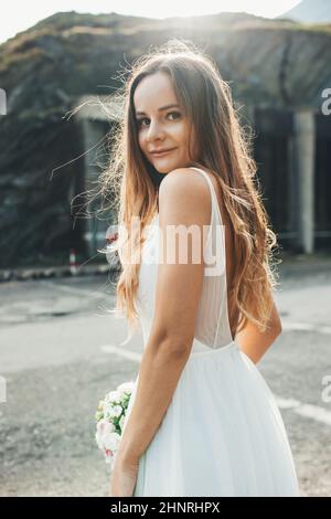 Portrait of a bride in love looking over her shoulder standing on the mountain road while she is at the photo shoot on her wedding day. Rustic Stock Photo