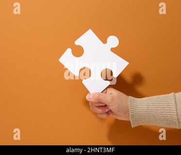female hand holds a fragment of a white large puzzle on a brown background, the concept of finding an idea, solving a problem Stock Photo