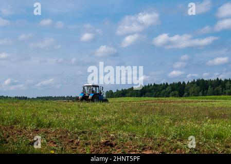 Tractor working in the field, plowing the land, preparing for sowing. View of the field and tractor in the distance against a blue sky with clouds Stock Photo