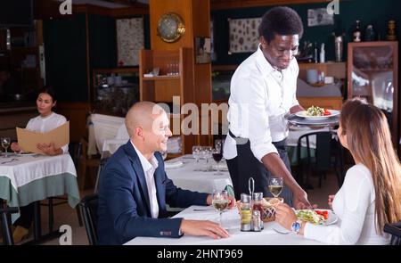 African American waiter bringing ordered dishes to couple Stock Photo