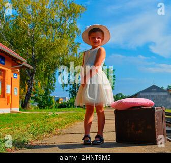 Adorable little girl on a railway station, waiting for the train with vintage suitcase. Traveling, holiday and chilhood concept. Travel insurance concept. Travel, tourism, summer vacation and family concept Stock Photo