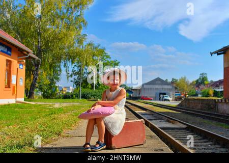 Adorable little girl on a railway station, waiting for the train with vintage suitcase. Traveling, holiday and chilhood concept. Travel insurance concept. Travel, tourism, summer vacation and family concept Stock Photo