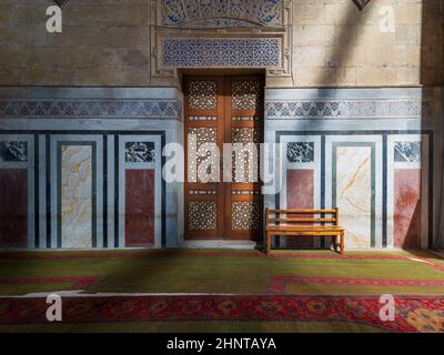 Ornamental wooden door, mosaic decorated walls, wooden bench, and floor lit by sunbeams, Al Rifai Mosque, Cairo, Egypt Stock Photo