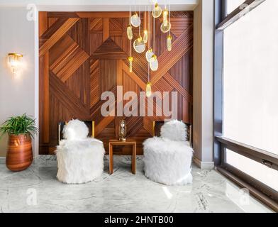 Two modern white feather armless chairs, small wooden modern table, plant pot, and decorated wood cladding wall Stock Photo