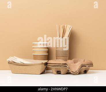 stack of brown disposable paper cups and a tray on a white table, wooden forks and knives, brown background. Utensils for takeaway drinks, zero waste Stock Photo