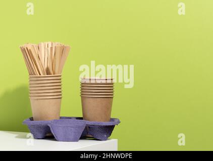 stack of brown disposable paper cups and a stand on a white table, green background. Utensils for takeaway drinks, zero waste Stock Photo