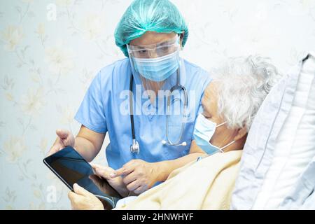 Asian doctor wearing face shield and PPE suit new normal holding tablet with senior woman patient protect safety infection Covid-19 Coronavirus outbre Stock Photo