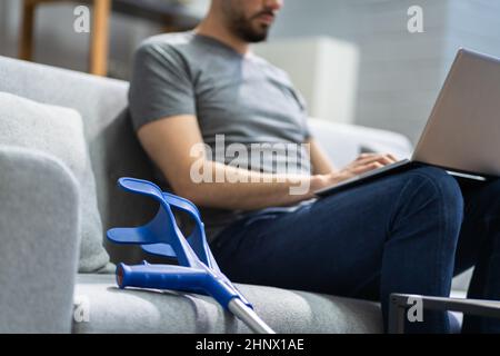 Injured Man With Broken Leg And Crutches Using Laptop Stock Photo