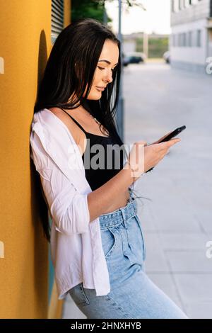 Woman using her mobile phone while leaning against a wall in the street. Stock Photo