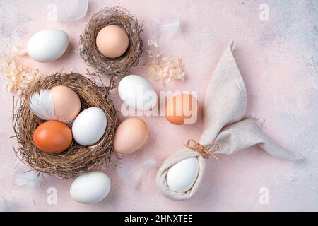 Happy Easter, festive background with natural eggs in the nest and an Easter bunny from a napkin on a light pink background. Top view, copy space. Stock Photo
