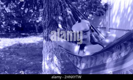 joyful woman wearing virtual reality glasses rests in a hammock near a tree, imagining a dream come true, an invented world. The concept of getting Stock Photo