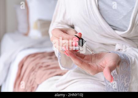 Cropped view woman in bathrobe applying serum on hands on bed in bedroom, close up. Unrecognizable lady keeping her skin moisturized using hyaluronic acid. Hand skin care concept Stock Photo