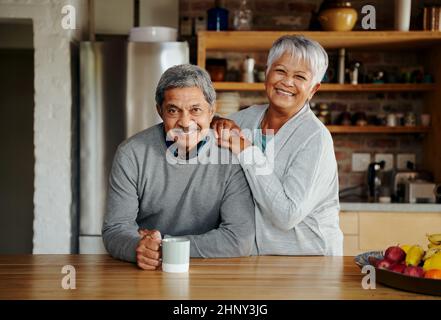 Portrait of happily retired elderly biracial couple standing, smiling at camera in their modern kitchen. Stock Photo