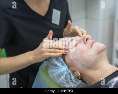 An adult woman of 63 years lies on the procedure of rejuvenation at the beautician with a mask on her face. Cosmetologist's hands massage the lobb. Hi Stock Photo