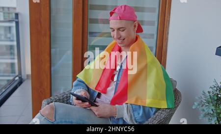 non binary person with pride rainbow flag on arms sitting on the balcony and looking at a smartphone with a smile. High quality photo Stock Photo