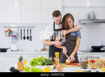 Smiling boyfriend hug woman from behind watching her preparing fruit salad in the kitchen, loving man embrace girlfriend cutting fresh fruit, romantic Stock Photo