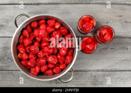 Table top view - homemade strawberry compote preparation. Metal pot full of strawberries, three bottles filled with syrup and fruits on the side, layi Stock Photo