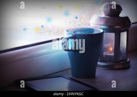 Winter decoration with lantern, hot tea cup,e-book reader and lights on window . Stock Photo