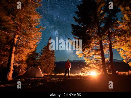 Back view of male camper standing on lawn between trees near tent and admiring surrounding beauty of starry sky with Milky way over snow-capped peaks of mountain hills. Night camping in the mountains. Stock Photo