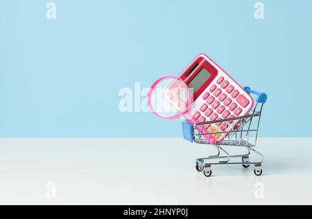 magnifier and pink plastic calculator in a miniature pink trolley on a blue background. Budget planning concept, savings counting, cost control Stock Photo