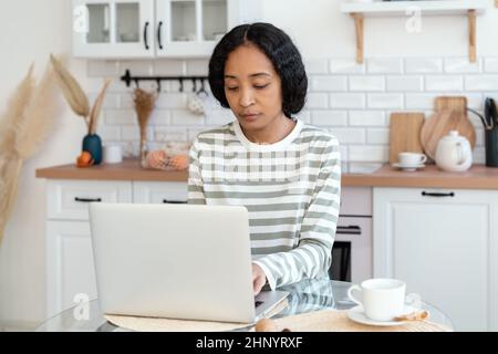 African-american female using laptop while sitting in kitchen. Confident and focused on remote work Stock Photo