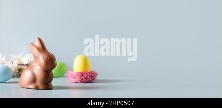 Easter banner with chocolate bunny and yellow egg, spring blossom flowers on blue background with copy space. Happy Easter greeting card. Stock Photo
