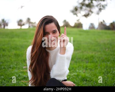 I have an idea. A young woman in the park looking at the camera smiling and raising her index finger. Stock Photo