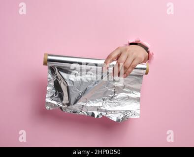 female hand holding a scroll of gray food foil on a pink background, part of the body sticking out of a torn hole Stock Photo