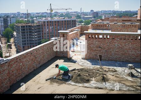 Construction site with newly built apartment buildings, tower crane and floor screeding contractor. Male worker using screed rail while screeding floor in building with brick walls. Stock Photo