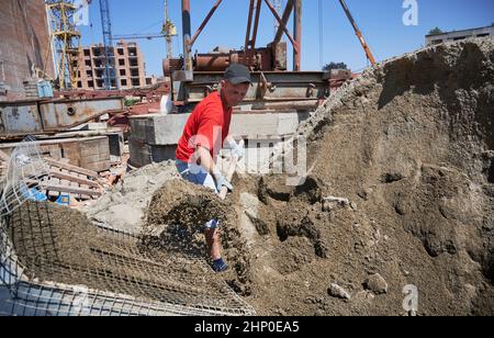 Male worker with shovel in his hands throwing cementitious floor screed material into container with net. Man contractor shoveling sand-cement mix outdoors at construction site. Stock Photo