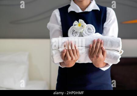 Closeup white towel with white flower on young hotel maid's hand. Staff in blue uniform preparing rooms for hotel guests. Stock Photo