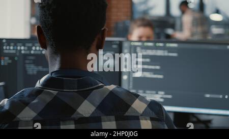 Portrait of african american programer online cloud computing on laptop sitting at desk with multiple screens parsing code. Over shoulder view of app developer working on user interface. Stock Photo