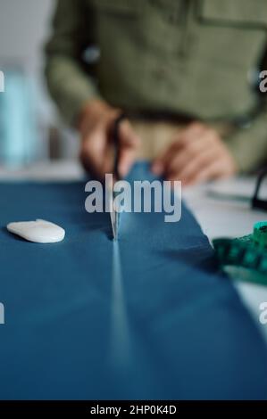 Dressmaker with scissors cuts cloth pattern at her workplace in workshop. Dressmaking occupation and professional sewing, handmade tailoring business Stock Photo
