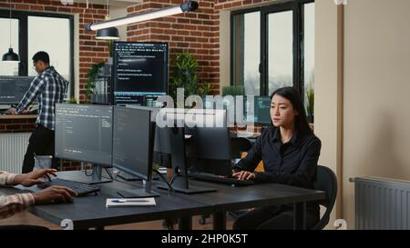 Programer with clipboard sitting down at desk in front of programer writing code while team of database admins compute big data. Team of mixed programmers working on machine learning software. Stock Photo