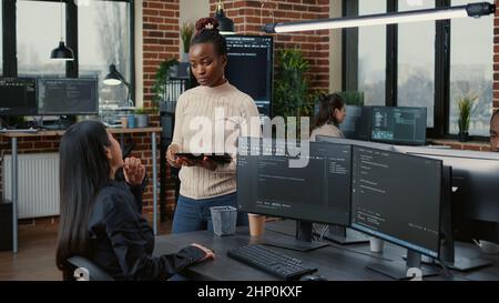 Software developer standing up holding digital tablet talking with programer sitting at desk with computer screens showing running code. System engineer coding with team writing machine learning data. Stock Photo