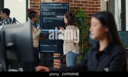 System engineers comparing source code on wall screen tv analyzing errors using digital tablet with focus switching to programer writing algorithm. Team of coders debugging machine learning data. Stock Photo