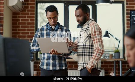 System engineers talking about algorithms holding laptop and standing in the middle of office with colleagues working. Team of software programmers discussing database looking at portable computer. Stock Photo