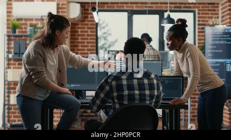 System engineer sitting on desk discussing with mixed team of coders about ai innovation in front of multiple screens compiling code. Programmers doing teamwork looking at machine learning algorithms. Stock Photo