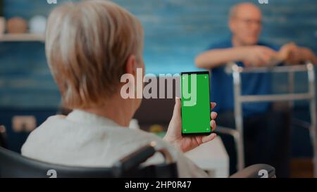 Senior woman in wheelchair holding smartphone with green screen at home. Retired person with chronic disability using isolated copy space with blank mock up and chroma key background on display. Stock Photo