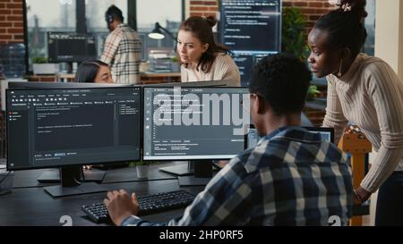 Mixed team of artificial intelligence cloud programers talking about programming in front of multiple screens compiling code. Programmers doing teamwork looking at machine learning algorithms. Stock Photo