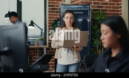 Portrait of caucasian app developer holding laptop looking up and smiling at camera. Software ai programmer typing on portable computer next to colleagues codingcyber security app. Stock Photo
