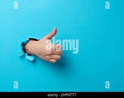 woman's hand sticks out of the torn hole and shows a gesture okay, thumb up. Blue background Stock Photo